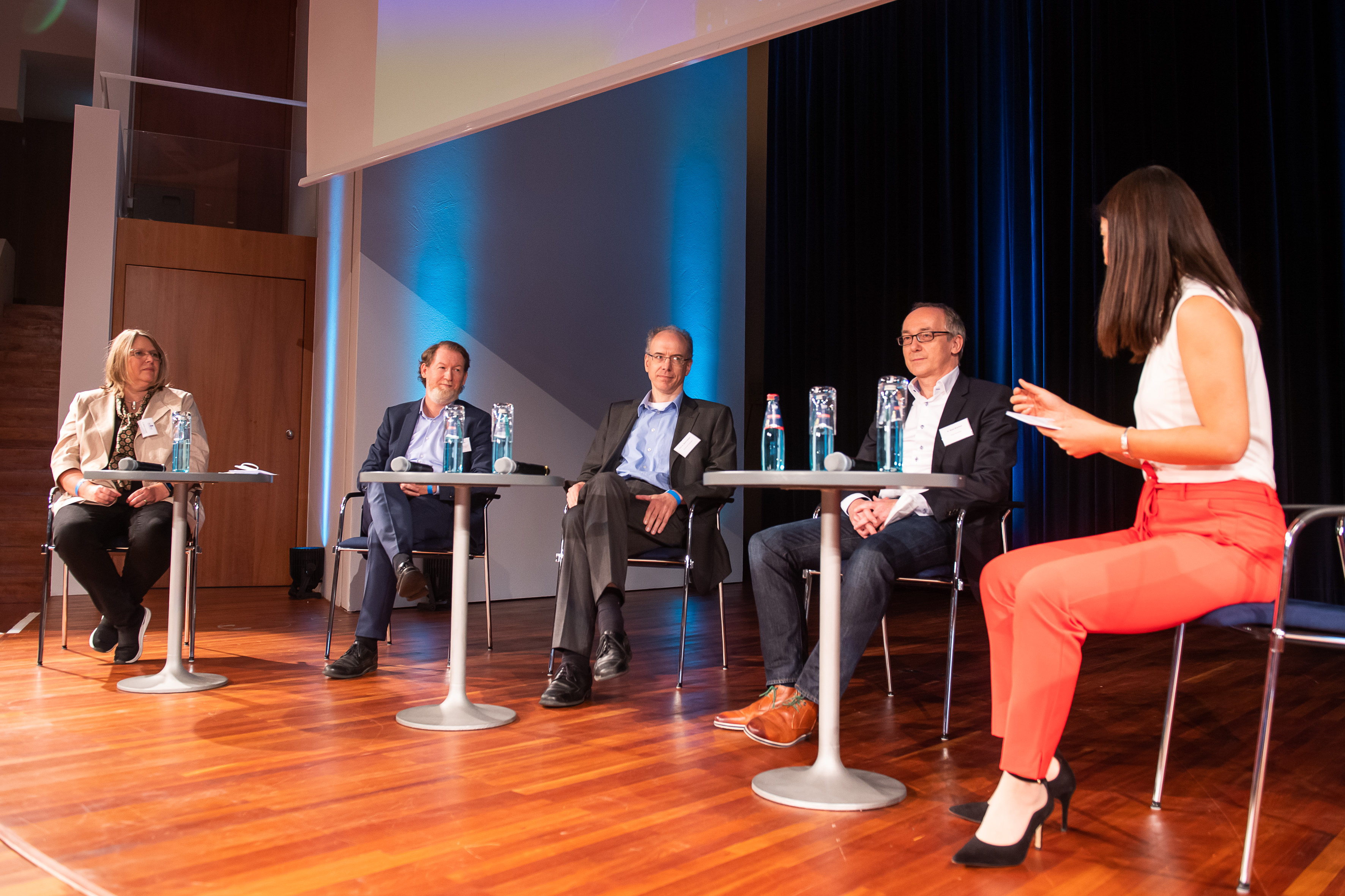 mehrere Personen sitzen an kleinen Tischen auf einer Bühne in einer Diskussionsrunde