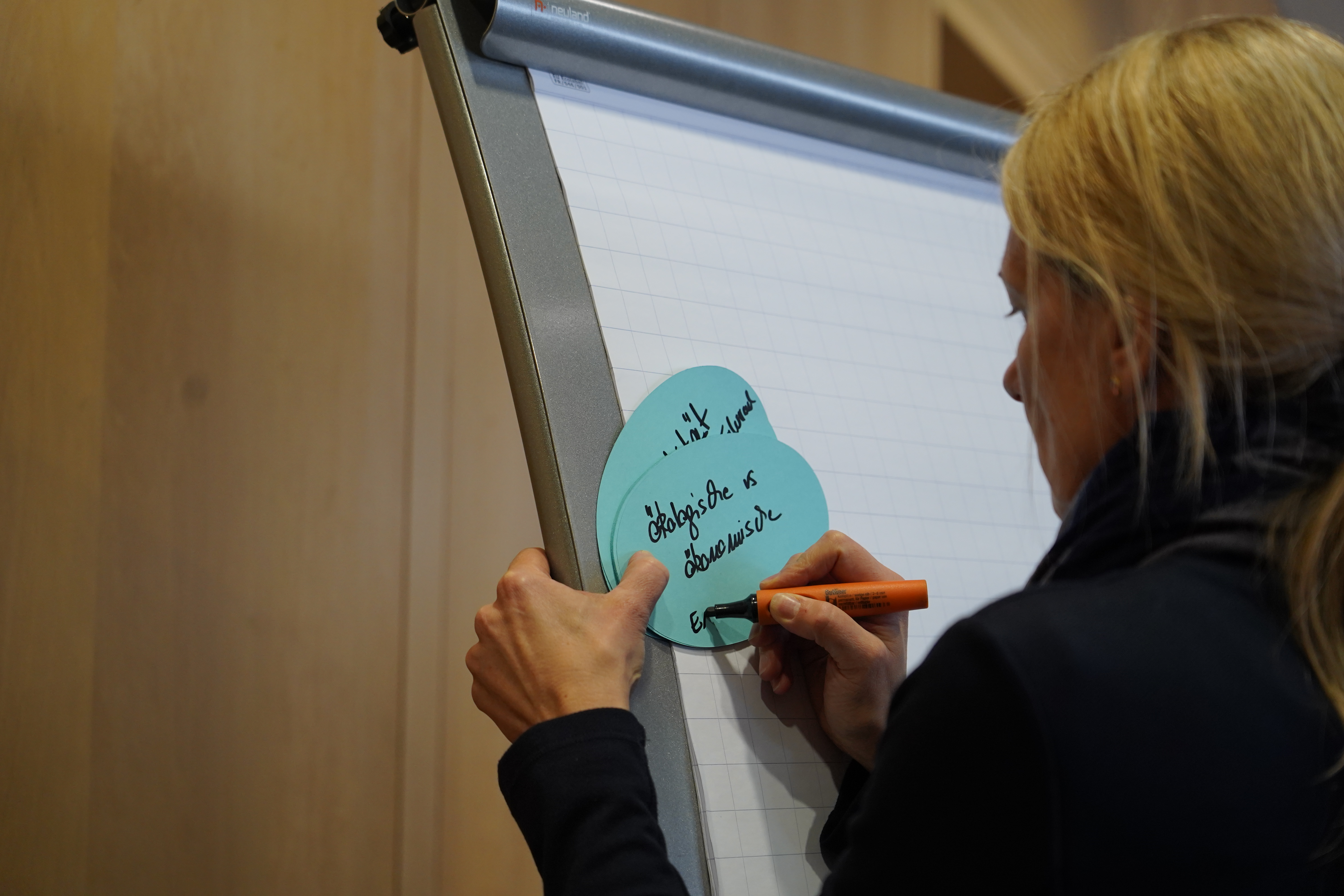 Eine Teilnehmerin schreibt im Rahmen des World Cafés auf eine hellblaue Karte die Worte "ökologisch vs. ökonomisch".