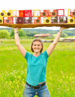 Die Firmenchefin auf einer blühenden Wiese, wie sie ein Brett mit vielen bunten Nudelpackungen stemmt.