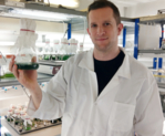 A man in a white lab coat holding in his hand an Erlenmeyer flask filled with a green liquid.